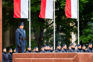 Grafika przedstawia sierżanta policji z bagnetem, w umundurowaniu galowym, trzy flagi Polski oraz ślubujących policjantów.