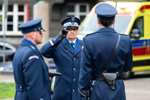 Na zdjęciu widoczny oddający honor Komendant Wojewódzki Policji w Katowicach oraz dwaj Policjanci na tle  żółtej karetki pogotowia ratunkowego