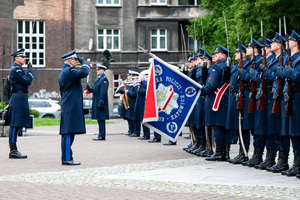 Grafika przedstawia oddającego honor Komendanta Wojewódzkiego Policji w Katowicach stojącego frontem do sztandaru Komendy Wojewódzkiej Policji w Katowicach. oraz kompanię honorową.