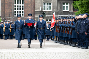 Na zdjęciu trzej policjanci w poczcie flagowym. Na tle kompanii honorowej, ślubujących policjantów  oraz orkiestry policyjnej.