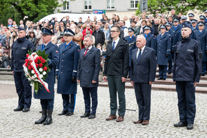 Na zdjęciu dwaj policjanci w mundurach galowych, dwaj policjanci w mundurach służbowych oraz inni zaproszeni goście. Policjant w mundurze galowym trzyma w ręku wieniec kwiatów.