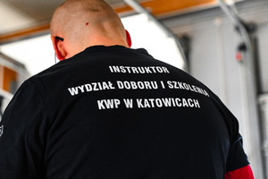 na zdjęciu policjant tyłem do obiektywu w koszulce z napisem instruktor wydziału doboru i szkolenia komendy wojewódzkiej policji w katowicach