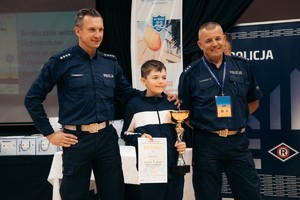 Na zdjęciu umundurowani policjanci pozujący do zdjęcia wraz z chłopcem trzymającym w ręku puchar.