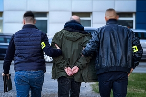 na zdjęciu policjanci po cywilu z opaskami na ramionach prowadzą zatrzymanego w kajdankach