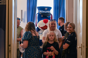 Zdjęcie przedstawia maskotkę Sznupka wraz z grupą dzieci oraz dwoma policjantami