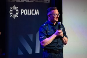 Zdjęcie przedstawia: umundurowanego policjanta podczas przemówienia z mikrofonem.