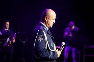 Zdjęcie przedstawia: policjanta z orkiestry z mikrofonem w dłoni.