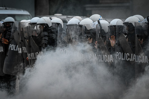 na zdjęciu policjant z tarczami w szyku przed nimi biały dym