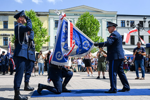 kolorowe zdjęcie przedstawiające policjanta trzymającego sztandar, przed którym klęczy policjant, w tle widać innych policjantów oraz osoby cywilne