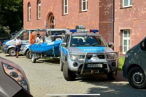 Kolorowe zdjęcie policjanta policji wodnej, w tle radiowóz z przyczepą, na której znajduje się łódź motorowa.