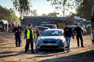 Zdjęcie przedstawia policjantów na miejscu zdarzenia wśród samochodów i kontenerów