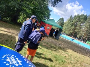 Na zdjęciu widoczny jest umundurowany funkcjonariusz Policji, który jest w towarzystwie dziecka. W tle widoczny jest namiot, pod którym znajduje się grupa młodzieży.