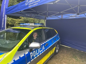 Na zdjęciu widoczny jest oznakowany radiowóz policyjny, za kierownicą którego znajduje się dziewczyna