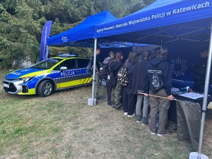 Na zdjęciu widoczny jest oznakowany radiowóz policyjny, a obok niego pod namiotem znajduje się grupa osób.
