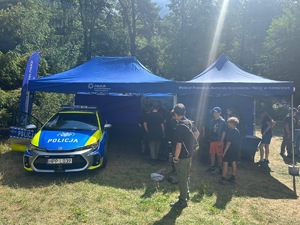 Na zdjęciu widnieją dwa namioty policyjne, pod którym znajduje się oznakowany radiowóz Policyjny wraz z grupą młodzieży.