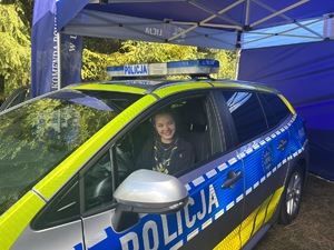 Na zdjęciu widoczny jest oznakowany radiowóz policyjny, za kierownicą którego znajduje się dziewczyna