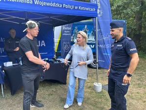 Na zdjęciu widoczne są trzy osoby, które ze sobą rozmawiają. W tle widoczny jest umundurowany funkcjonariusz Policji, który stoi przed stoiskiem z wyposażeniem Policji.
