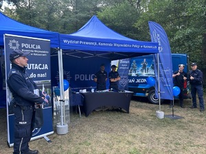 Na zdjęciu widoczne jest stoisko policyjne, za którym stoi dwóch funkcjonariuszy Policji. Obok stoiska znajduje się kolejne trzy osoby, z których dwóch to policjanci.