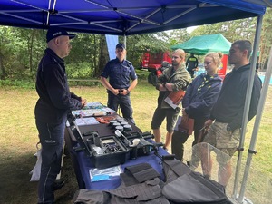 Na zdjęciu widoczne jest policyjne stoisko z policyjnym wyposażeniem, któremu przygląda się grupa osób.