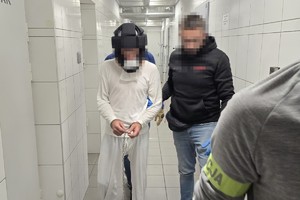 na zdjęciu widać podejrzanego ubranego w  dres koloru białego, z założonymi kajdankami zespolonymi na nogi i ręce który jest prowadzony przez nieumundurowanych policjantów przez korytarz