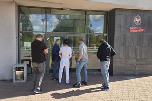 na zdjęciu widać doprowadzonego przez policjantów podejrzanego mężczyznę ubranego w dres koloru białego z założonymi kajdankami zespolonymi na nogi i ręce przed wejście do budynku Prokuratury  Rejonowej w Dąbrowie Górniczej