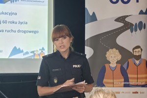 Na zdjęciu widoczny jest umundurowany funkcjonariusz Policji, który prowadzi akcję informacyjno-edukacyjną związaną z bezpieczeństwem seniorów a w jego tle znajduje się plakat.