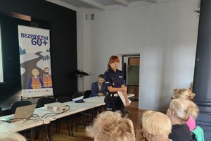 Na zdjęciu widoczny jest umundurowany funkcjonariusz Policji, który prowadzi akcję informacyjno-edukacyjną związaną z bezpieczeństwem seniorów. Obok policjantki znajdują się siedzący seniorzy.