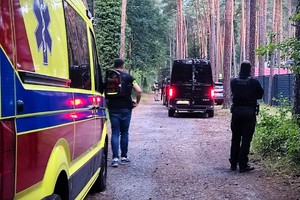 Zdjęcie przedstawia karetkę stojącą w zalesionym terenie oraz nieumundurowanych policjantów.