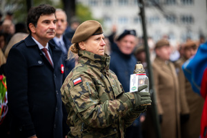 żołnierka stojąca obok cywili i trzymająca w ręku zapalony znicz