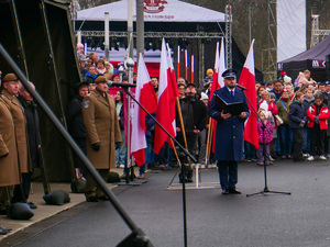 policjant przemawia przez mikrofon na tle cywilów oraz flag Polski