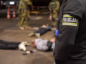 zdjęcie przedstawia rękę funkcjonariusza Policji, w drugim planie leżące na ziemi osoby.