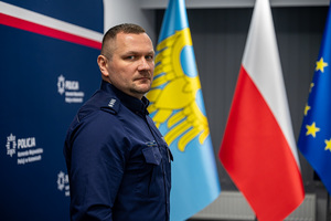 Policjant na tle niebieskiej ściany z napisem "Komenda Wojewódzka Policji w Katowicach" oraz flag.