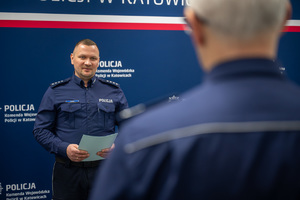 Dwaj policjanci zwróceni do siebie na tle niebieskiej ściany z napisem "Komenda Wojewódzka Policji w Katowicach".