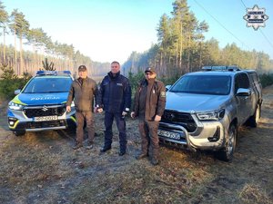 Policjant i dwóch funkcjonariuszy Straży Leśnej stojący przed radiowozem oraz pojazdem Straży Leśnej.