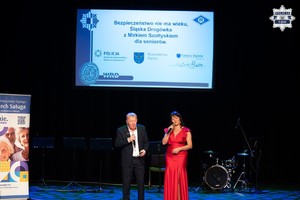 Elegancko ubrani kobieta i mężczyzna stoją na scenie z mikrofonami w dłoniach.