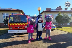 Maskotki Policji oraz Tauron stoją przed budynkiem na tle radiowozu i wozu Straży Pożarnej.