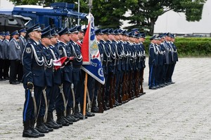 na zdjęciu umundurowani policjanci w mundurach galowych ze sztandarem stoją na baczność w szyku na placu