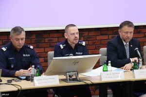 Panowie Komendant Główny Policji, Zastępca Komendanta Głównego Policji oraz Minister Spraw Wewnętrznych i Administracji siedzą przy biurku przed ekranem komputera i mikrofonami.