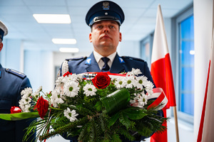 Zbliżenie na policjanta niosącego w dłoniach wieniec z kwiatów.