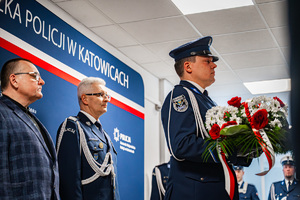 Policjant w mundurze galowym oraz elegancko ubrany mężczyzna stoją za plecami policjanta trzymającego w dłoniach wieniec z kwiatów.