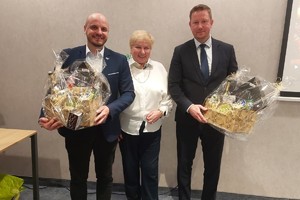 Dwaj mężczyźni z koszami prezentowymi  stoją w towarzystwie kobiety.