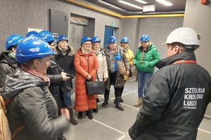 Grupa kobiet w niebieskich kaskach ochronnych stoi przed mężczyzną w białym kasku.