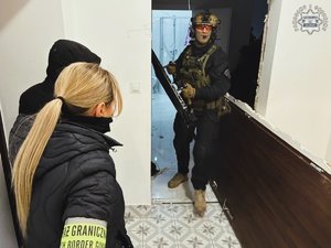 Na zdjęciu widać policjanta z taranem w ręku, obok wyważone drzwi. Na pierwszym planie funkcjonariusze straży granicznej.