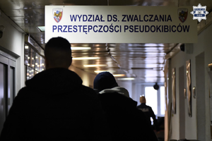 Na zdjęciu widać mężczyzn idących korytarzem, który oznaczony jest napisem: Wydział ds. zwalczania przestępczości pseudokibiców.