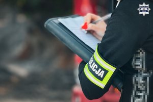 Zdjęcie przedstawia policjanta operacyjnego z opaską z napisem Policja, który wypełnia dokumentacje długopisem