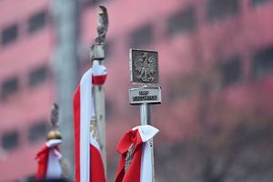 Zdjęcie przedstawia wierzchołek sztandaru z którego zwisa białoczerwona szarfa. Sam szczyt zakończony jest godłem narodowym (orzeł z koroną), a pod nim tabliczka z napisem KWP (Komenda Wojewódzka Policji) Katowice