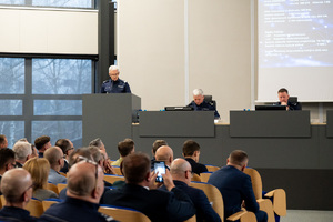 Kolorowe zdjęcie przemawiającego Generała, obok widoczni Jego zastępcy. W tle widoczna widownia siedząca na krzesłach rotundy.