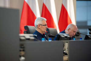 Kolorowe zdjęcie Generała na tle jego zastępcy siedzących za mównicą na tle flag Polski.