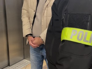 Zbliżenie na opaskę z napisem Policja na ramieniu policjanta i w tle stojącego zatrzymanego zakutego w kajdanki