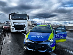 Na zdjęciu czerwony samochód osobowy przygnieciony do barierki drogowej przez białą ciężarówkę oraz radiowóz